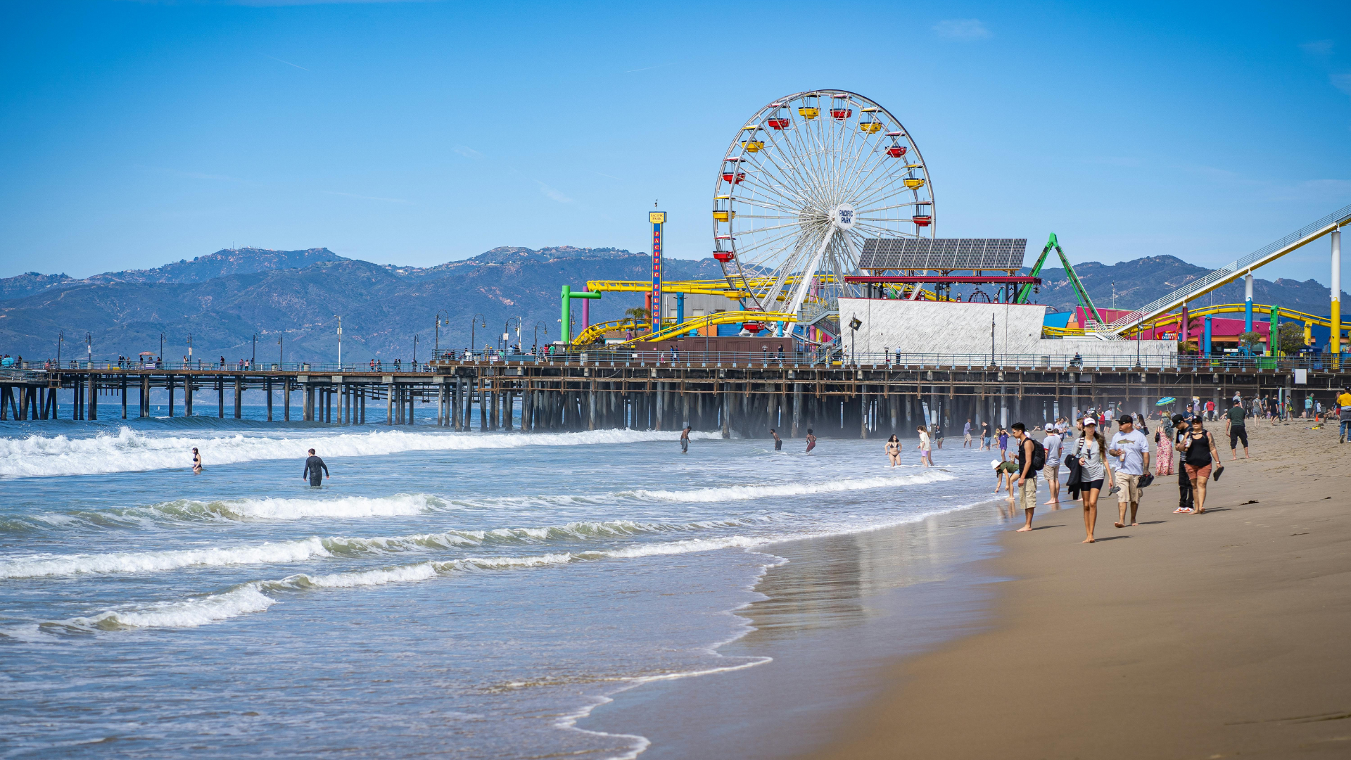 People stroll on a sandy beach with a vibrant Ferris wheel nearby, capturing a fun day at Santa Cruz.