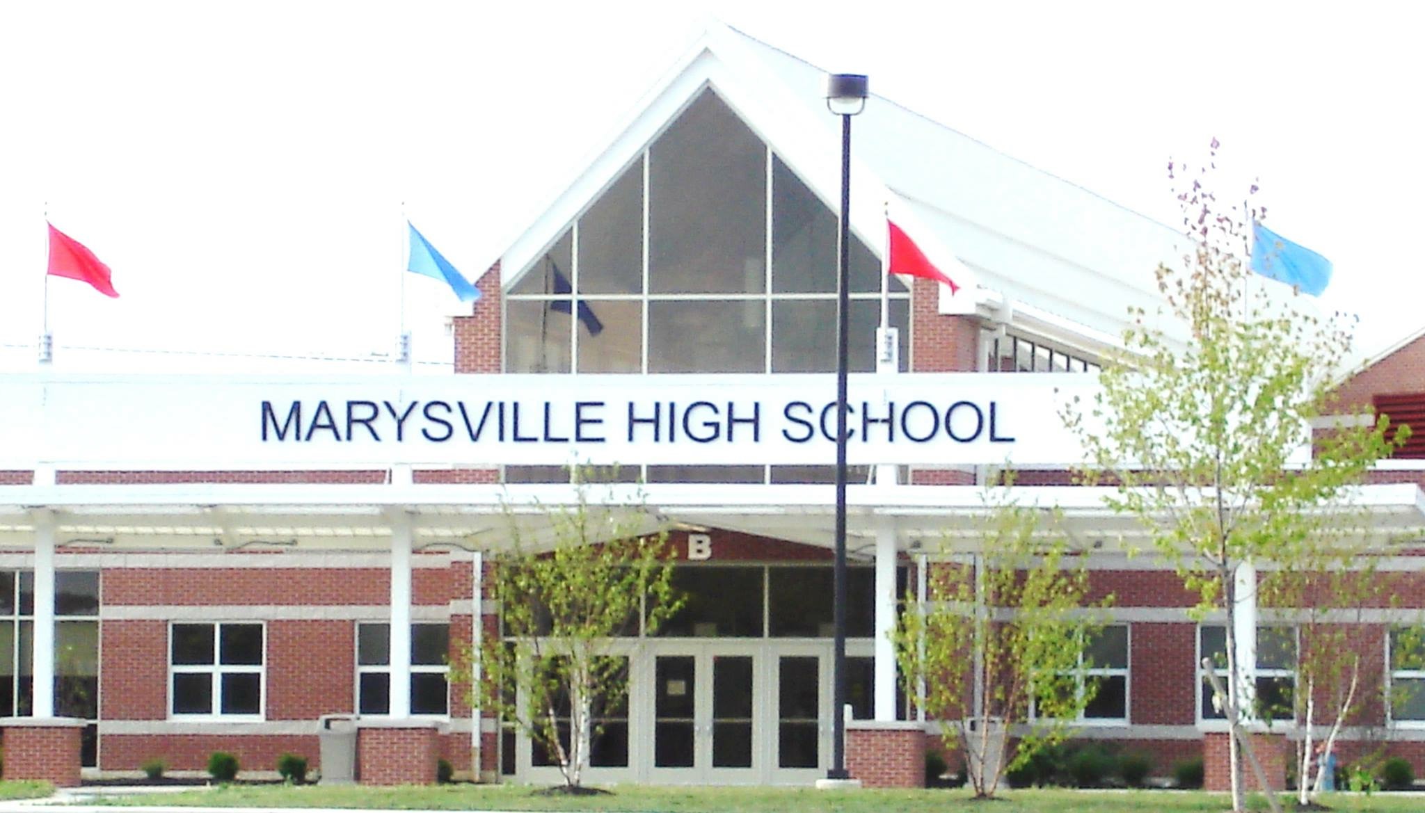 Front entrance of Marysville High School in Marysville, Ohio, with brick exterior, large glass windows, and flags flying above the sign