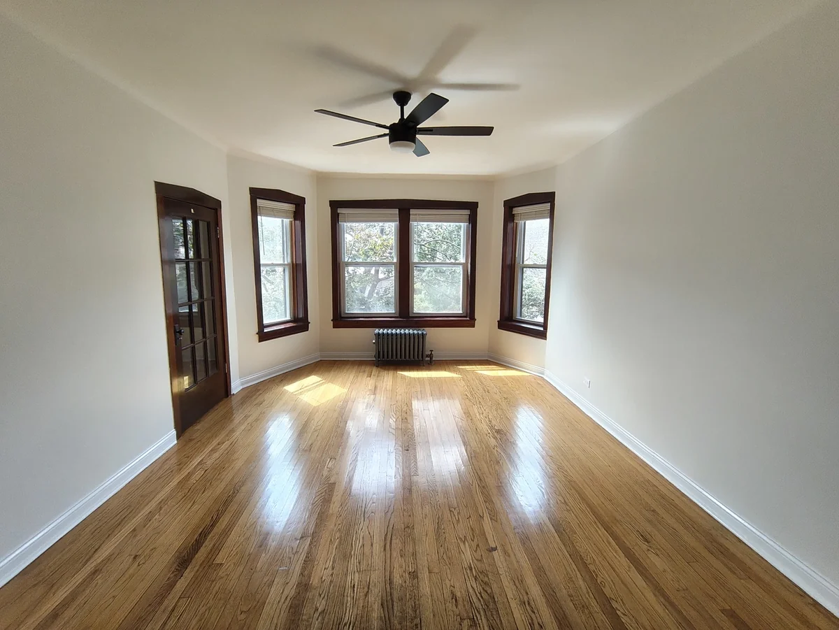 Empty living room with hardwood floors and a bank of three windows in a renovated Rogers Park 2-bedroom apartment