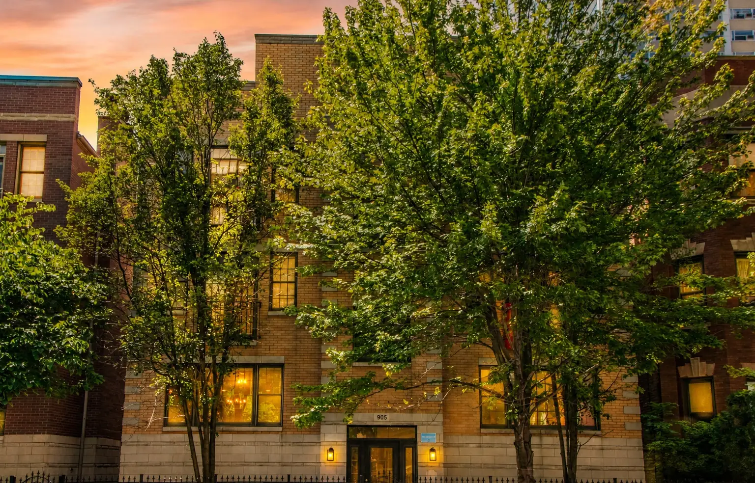 Tan brick three-story building at 905 W Lawrence Ave in Uptown, Chicago, behind leafy street trees at dusk