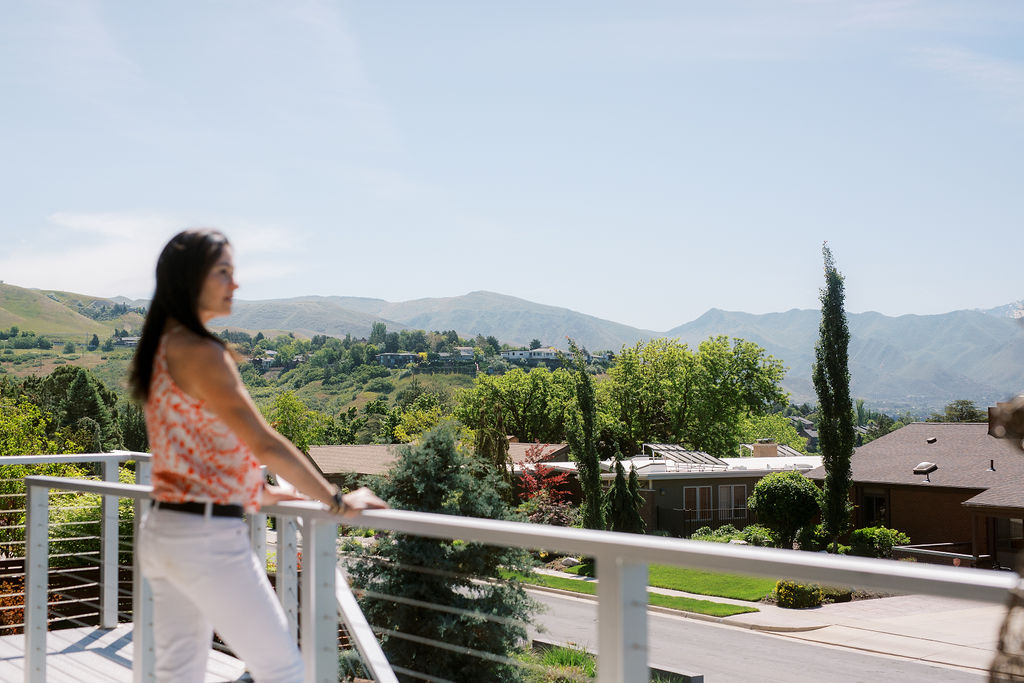 a woman with dark hair stands at a balcony and looks at the mountains beyond