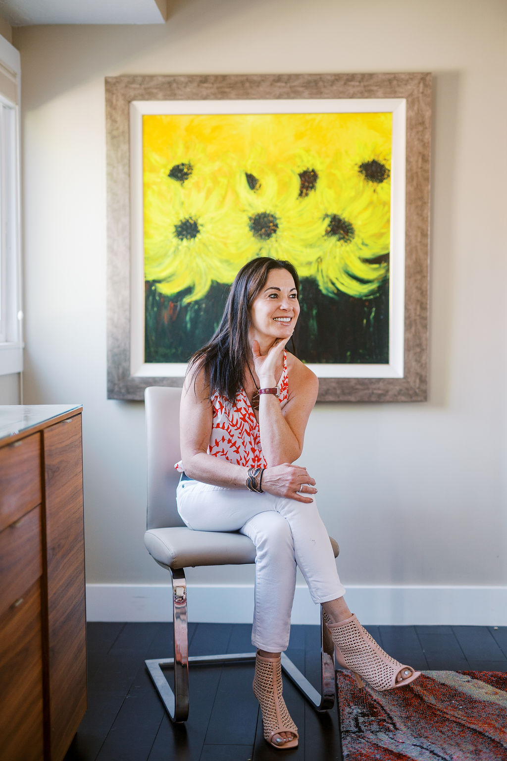 a woman with dark hair and white pants smiles in front of a painting of sunflowers