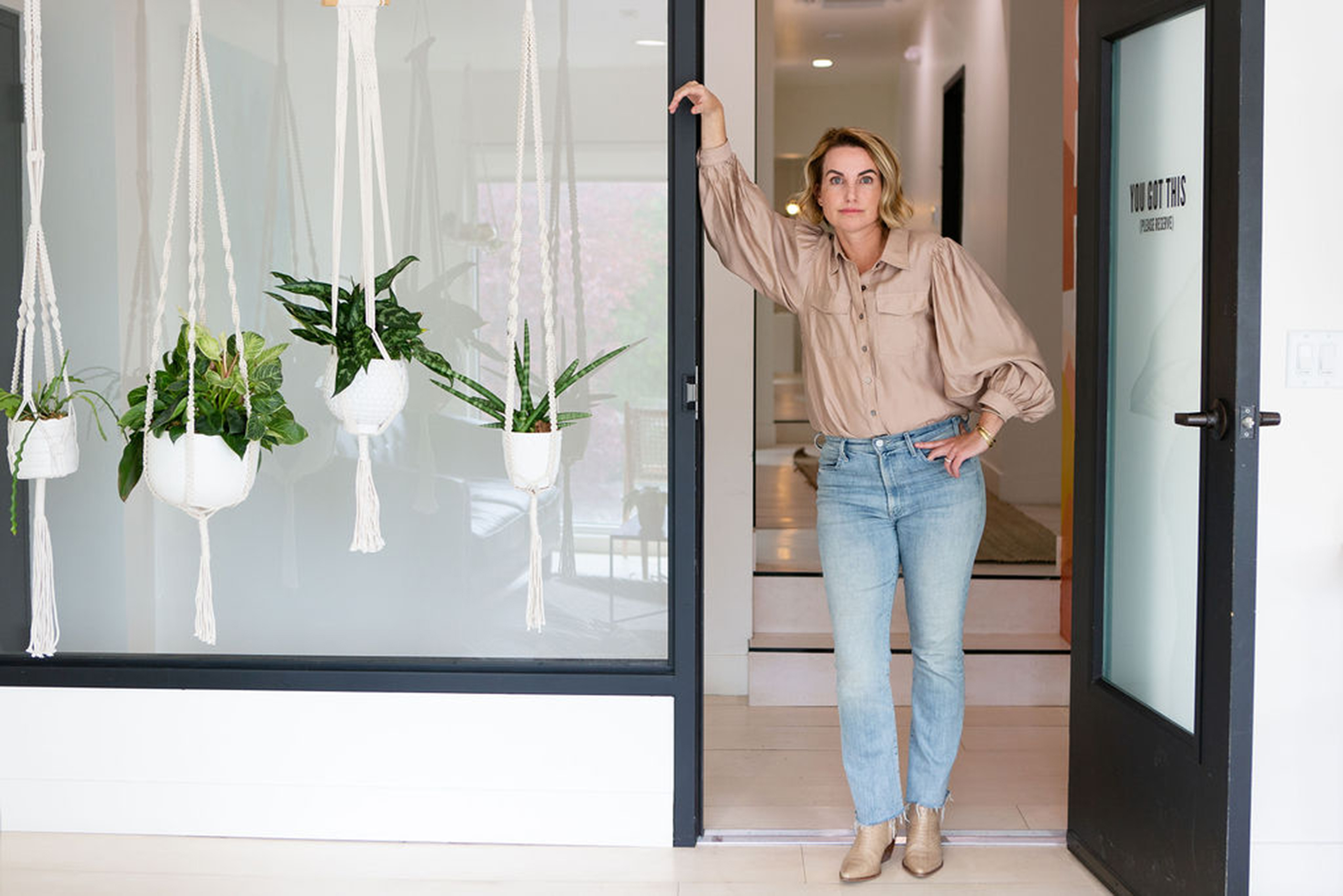 an attractive middle aged blonde woman stands in the doorway of an inviting office space with a glass door and hanging house plants